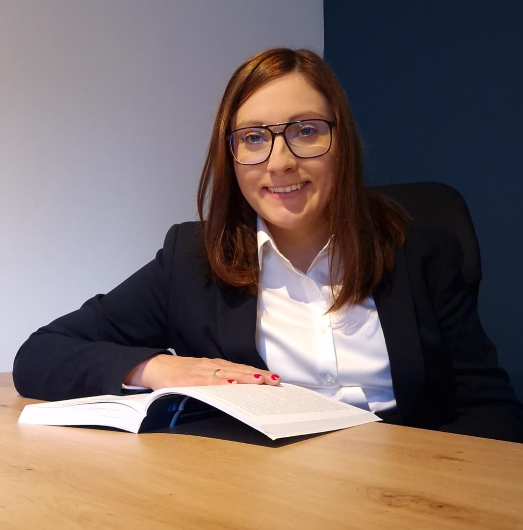 Młoda kobieta w okularach w kwadratowych oprawkach, uśmiechnięta, siedząca przy drewnianym biurku w biurze z szarym i niebieskim tłem. Ubrana w ciemny garnitur i białą koszulę, trzyma otwartą książkę w lewej ręce, pozując profesjonalnie.