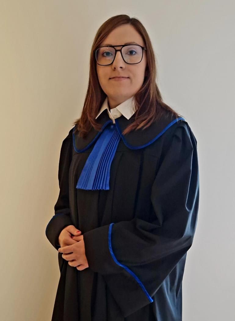 Młoda kobieta w okularach w kwadratowych oprawkach, ubrana w czarną togę prawniczą z niebieskimi wykończeniami i białym kołnierzykiem, stojąca z rękami splecionymi przed sobą na białym tle.